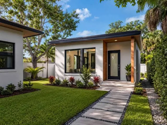 Modern single-story accessory dwelling unit with white stucco walls, a wood-trimmed entry, large windows and a paver walkway through a landscaped yard