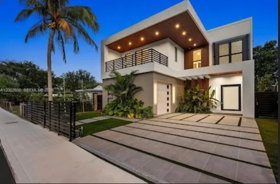 Contemporary two-story home at dusk with a wood-clad soffit, a balcony with black railing, an oversized paver driveway and a palm tree