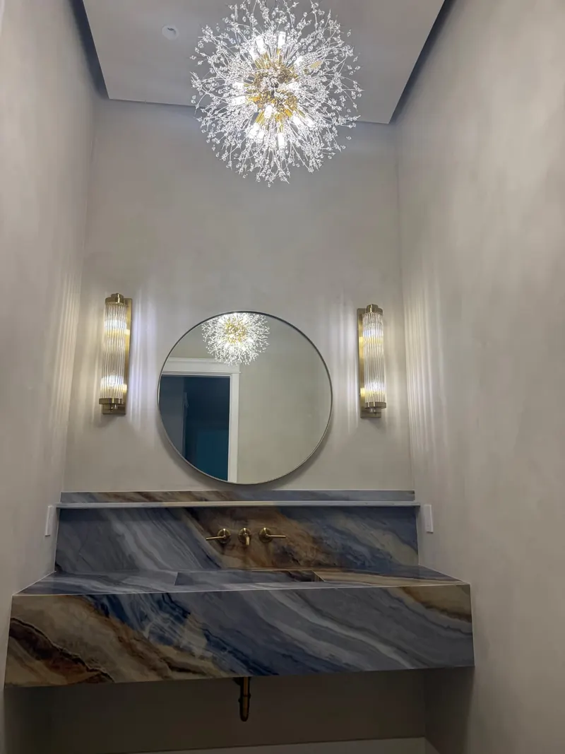 Powder bathroom with a blue and gold stone vanity top, round mirror, wall sconces and a spherical chandelier