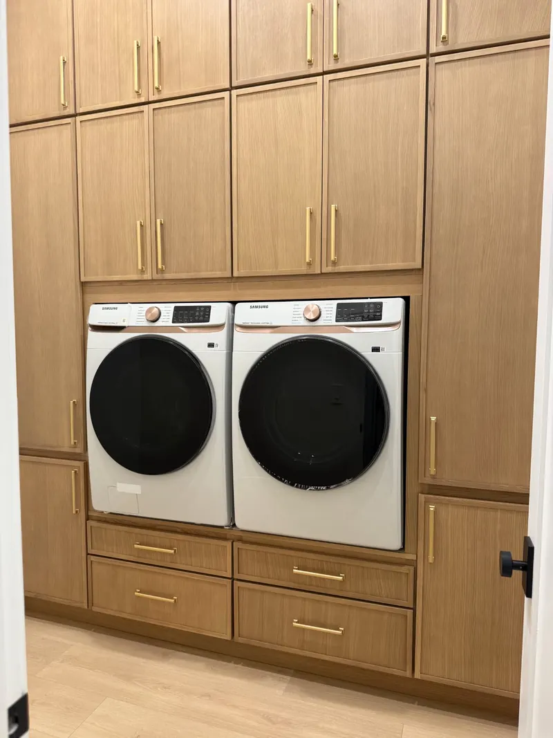 Laundry room with side-by-side washer and dryer, wood-grain cabinets to the ceiling and brass handles
