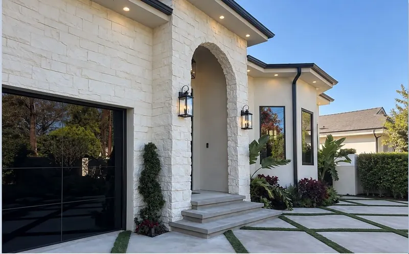 Cream stone house with an arched entry, steps, wall lanterns and a black glass garage door